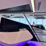 “Glossy white curves + purple glow + mirror drama = office entrance on another level.” A futuristic high-rise reception desk in glossy white with flowing organic curves and subtle purple-blue LED underlighting. Behind it, dramatic angular black-and-white geometric partitions with mirrored sections reflect the office, while a panoramic city view is visible through floor-to-ceiling windows.