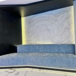“Powder-blue velvet + glowing marble waves = the ultimate chill pod.” A futuristic built-in seating nook with a long, low light-blue upholstered bench set into a curved matte-black alcove. The back wall features an illuminated abstract wavy marble panel with warm hidden LED backlighting and subtle edge glow under the bench.