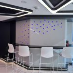 “Blue LED triangle rain + floating counter glow = morning coffee just got futuristic.” A futuristic high-end home bar or coffee station with a curved matte light-gray counter outlined in warm LED glow strips. Four modern white bar stools with chrome legs are positioned in front. The back wall features an artistic scattering of small illuminated blue triangle lights in a cascading pattern, set against black accents and glossy white marble flooring.