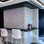 “Blue triangle LED rain + black counter + hidden glow = bar nook goals unlocked.” A sleek, futuristic home bar nook with a curved black countertop and matte light-gray paneling outlined in warm LED strips. Two modern white bar stools with metal legs sit in front. The back wall features an artistic arrangement of small illuminated blue triangle lights forming an abstract cascading pattern.