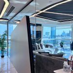 “Curved gray cloud sofa + that insane city view = penthouse perfection unlocked.” A luxurious high-rise open-plan living room with panoramic city views through floor-to-ceiling glass walls. A plush curved gray sectional sofa faces a marble coffee table, partially separated by a sleek angular matte-black room divider. Modern curved LED ceiling lights and glossy white marble flooring complete the ultra-contemporary scene.