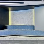 “Powder-blue velvet + glowing marble waves = quiet luxury went full cyberpunk.” A futuristic built-in seating nook with a long, low light-blue upholstered bench set into a curved black alcove. The back wall features an illuminated abstract wavy marble or stone panel with subtle LED glow, edged by dramatic hidden lighting under the bench and along the ceiling.