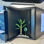 “Black pod + glowing monstera = the most extra plant nook in existence.” A futuristic black matte alcove with organic curved walls featuring a built-in illuminated planter containing a large artificial banana leaf plant and a monstera, surrounded by white pebbles. The niche is dramatically lit from above, set against glossy white marble flooring in a high-tech luxury interior.
