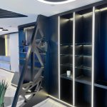 “Matte black + hidden LEDs + laser-cut drama = quiet luxury after dark.” A sleek modern built-in shelving unit in matte black with integrated warm LED strip lighting between each bay, featuring four open glass shelves on the right and a dramatic geometric laser-cut black metal partition on the left. The unit is set against a curved ceiling with hidden ambient lighting, on glossy white marble flooring.