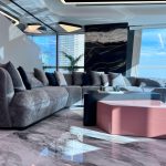 “Curved gray cloud sofa + black marble wall + that pop of pink = penthouse goals unlocked.” A luxurious high-rise living room with floor-to-ceiling windows showcasing a city skyline. A large curved gray plush sectional sofa faces a modern two-tier coffee table in matte black and glossy pink. A dramatic black-and-gold marble feature wall, potted plants, and geometric pendant lights complete the ultra-modern scene on glossy marble flooring.