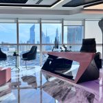 “Pink lacquer desk + Dubai skyline = closing million-dollar deals in style.” A futuristic high-rise executive office with panoramic Dubai skyline views (including the Burj Khalifa) through floor-to-ceiling windows. A dramatic angular desk in glossy pink-and-black lacquer dominates the space, paired with a high-back black leather chair and two curved gray visitor chairs on a polished marble floor.