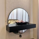 A sculptural mirror and floral accents bring elegance to this minimalist bedroom console setup. Bedroom or dressing area with circular black-framed mirror, black floating shelf, decorative bowl of flowers, and light beige tiled flooring.