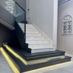Floating stone steps and ambient lighting define this sleek staircase, where modern design meets architectural finesse. Indoor staircase with light stone steps, black base platform with yellow LED lighting, and glass railing in a modern minimalist interior.