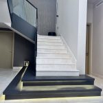 Sleek marble steps and ambient lighting define this modern staircase, where minimalism meets architectural elegance. Modern staircase with white marble steps, glass railing, and black platform base with integrated lighting in a minimalist interior.