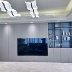 A sleek media wall and sculptural lighting elevate this minimalist living space with refined elegance. Modern living room with wall-mounted TV, built-in shelving, wavy ceiling lights, and a neutral palette of gray and beige.