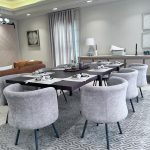 “Gray barrel chairs + bubble chandelier + all the natural light = dining room goals unlocked.” A bright modern open-plan dining room with a long dark-wood rectangular table set for eight, surrounded by plush light-gray upholstered barrel chairs on black metal legs. The table features gray placemats, white plates, and minimalist tableware under a contemporary bubble-glass chandelier. A large geometric gray area rug sits beneath, with floor-to-ceiling sheer white curtains, a cognac leather sofa visible on the left, and neutral abstract artwork on the walls.