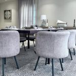 “Gray barrel chairs + bubble chandelier + all the natural light = dining goals unlocked.” A bright modern dining room with a long dark-wood rectangular table surrounded by eight plush light-gray upholstered barrel chairs on black metal legs. The table is elegantly set for a meal with gray placemats, white plates, and glassware under a contemporary bubble-glass chandelier. A large geometric gray area rug sits beneath, with floor-to-ceiling sheer white curtains, a cognac leather sofa visible on the left, and minimalist white artwork on the walls.