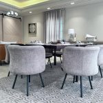 “Gray barrel chairs + bubble chandelier + all the natural light = dining goals unlocked.” A bright modern dining room with a long dark-wood rectangular table surrounded by eight plush light-gray upholstered barrel chairs on black metal legs. The table is elegantly set for a meal with gray placemats, white plates, and glassware under a contemporary bubble-glass chandelier. A large geometric gray area rug sits beneath, with floor-to-ceiling sheer white curtains, a cognac leather sofa visible on the left, and minimalist white artwork on the walls.