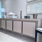 “When your storage is this pretty, clutter simply isn’t invited.” A long, minimalist light-oak sideboard with four seamless doors and subtle black trim, styled with a soft blue-gray ceramic lamp, white abstract sculpture vase with dried stems, woven basket with folded linens, black twisted candlestick, teal mugs on a tray, and a beige planter. Two large framed white textured circular artworks hang above, set against a neutral wall with sheer curtains and glossy light marble flooring.