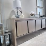 “When your storage looks this good, clutter doesn’t stand a chance.” A sleek modern light-oak sideboard with four matte doors and recessed black handles, styled with a blue-gray ceramic table lamp, white abstract sculpture vase, woven basket with tissues, and black candle holders. A small round gray side table in the foreground holds textured candles, all on glossy light marble flooring near sheer curtains.