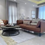 “When your sofa is this buttery, staying in becomes the only plan.” A sunlit modern open-plan living room featuring a cognac-brown leather three-seater sofa and matching armchair arranged around a matte-black oval two-tier coffee table on a light geometric area rug. Tall sheer white curtains with soft gray drapes frame large windows, a white sideboard displays minimalist decor, and neutral abstract artwork hangs on the walls.