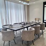“Taupe barrel chairs + bubble chandelier + all the natural light = dining goals unlocked.” A bright modern dining room with a long rectangular black dining table set for eight, surrounded by plush taupe-gray upholstered barrel chairs with black metal legs. The table is styled with gray placemats, white plates, and minimalist tableware under a contemporary bubble-glass chandelier. Floor-to-ceiling sheer white curtains frame tall windows, and a large geometric gray area rug sits beneath on light marble flooring.