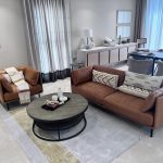 “When your sofas are this buttery, canceling plans becomes a lifestyle.” A bright, modern open-plan living-dining room featuring a cognac-brown leather three-seater sofa and matching armchair arranged around a round black metal coffee table on a light geometric area rug. Tall sheer white curtains with gray drapes frame large windows, a white sideboard with decor sits in the background, and a contemporary dining area with gray upholstered chairs is visible.