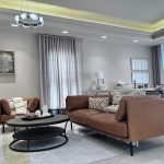 “When your sofas are this buttery, shoes might be optional.” A bright, modern open-plan living room with two cognac-brown leather sofas facing each other around a black oval two-tier coffee table on a light geometric area rug. Tall sheer white curtains frame large windows, a contemporary circular chandelier with glass globes hangs overhead, and neutral abstract artwork adorns the walls.