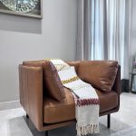 “When one chair steals the entire room’s attention.” A luxurious cognac-brown leather accent armchair with slim black metal legs, positioned in a bright modern corner. A textured cream throw blanket with mustard and burgundy stripes is casually draped over one arm and seat, next to tall light-gray curtains and a large abstract metallic wall clock above.