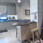 “When your kitchen peninsula is smoother than your weekend plans.” A sleek modern kitchen corner featuring matte gray cabinetry, a curved wooden breakfast bar peninsula with light oak base and dark countertop, and two wooden bar stools. Under-cabinet lighting illuminates the backsplash, with a single sculptural black pendant light overhead and marble-effect wall cladding on the right.