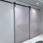 “Taupe geometric herringbone barn doors just invented closet drama.” A pair of oversized modern sliding barn doors in soft taupe-gray with geometric diagonal herringbone/Chevron paneling, mounted on a matte-black industrial track. The double doors are shown closed, covering a full wall in a minimalist interior with light flooring and recessed ceiling lighting.
