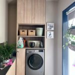 “Light oak + woven baskets + zero clutter = laundry room goals unlocked.” A compact, modern laundry nook featuring a front-load silver washing machine built into light oak cabinetry with upper storage cabinets, open shelves holding woven baskets and laundry supplies, and two framed motivational prints on the wall. A large window on the right brings in natural light, with hanging plants visible outside.