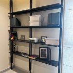 “When your shelving unit is basically a trophy case for winning at life.” A modern matte-black metal wall-mounted shelving unit with four tiers of open shelves displaying organized storage boxes, binders, books, framed awards, and decorative items against a light beige wall in a home office or study.