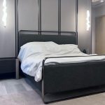 “When your headboard wall is basically architectural art.” A sleek modern bedroom featuring a king-size bed with a charcoal-gray upholstered frame and tall headboard accented by thin metallic trim. Crisp white bedding is neatly made, flanked by a minimalist black nightstand on the left. Two sculptural wavy pendant lights in white hang asymmetrically above, set against a dramatic paneled gray wall with vertical black metal strips and warm hidden LED lighting.
