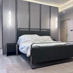 “When your headboard wall is basically architectural art.” A sleek modern bedroom featuring a king-size bed with a charcoal-gray upholstered frame and tall headboard accented by thin metallic trim. Crisp white bedding is neatly made, flanked by a minimalist black nightstand on the left. Two sculptural wavy pendant lights in white hang asymmetrically above, set against a dramatic paneled gray wall with vertical black metal strips and warm hidden LED lighting.