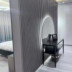 “When your console nook looks calmer than a luxury spa.” A modern entryway or dressing corner featuring a large arched frameless mirror with soft LED backlighting creating a glowing halo. A slim matte-black metal console table below holds two small framed photos and black candle holders, with a woven basket on the lower shelf. The backdrop is a dramatic dark-gray vertical fluted wood slat accent wall, on light wood-look flooring.