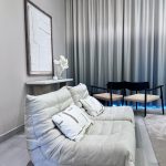 “When your lounge area looks calmer than a spa waiting room.” A serene modern lounge corner featuring a low, plush cream Togo-style sofa with deep tufted cushioning and two neutral throw pillows. Two minimalist black wooden armchairs with woven seats sit nearby, against floor-to-ceiling gradient gray-to-white ombre curtains. A small marble console holds a white floral arrangement and abstract wall art above, all on light gray flooring.