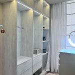 “Light wood + hidden LEDs = walk-in wardrobe perfection.” A bright, modern walk-in closet with floor-to-ceiling light wood-grain cabinetry featuring open shelving, hanging rods, and lower drawers. Warm integrated LED strip lighting runs along the top of each section, illuminating neatly organized clothes, shoes, and accessories. A large window with sheer white curtains is visible on the right.