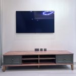 “When your TV console is prettier than most coffee tables.” A wall-mounted flat-screen TV above a long mid-century modern TV console in warm walnut wood with two sage-green drawer fronts featuring brass ring pulls. The unit has open center shelving and sits on tapered wooden legs against a plain white wall, on light gray wood-look flooring.