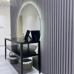 “When your console is minimalist but the mirror does all the talking.” A minimalist modern entryway console featuring a sleek black metal table with a lower shelf holding two woven black-and-white baskets. Above it, a large frameless arched mirror with integrated warm LED backlighting creates a glowing halo effect, set against a dramatic vertical gray fluted wood slat wall.