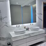 “Double vessel sinks + marble everything + floating vanity = quiet luxury bathroom goals.” A luxurious modern double vanity bathroom with a large frameless backlit mirror featuring a vertical blue LED accent strip in the center. Two oval white vessel sinks sit on a floating marble-effect countertop with dramatic gray veining, above a light wood-grain vanity with six soft-close drawers. The walls are clad in matching large-format marble porcelain tiles.