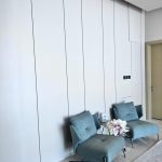 “Seamless white wall + dusty teal velvet = the definition of quiet luxury. Two modern teal-blue upholstered lounge chairs with slim black metal legs positioned against a seamless tall white paneled wall. A small round white marble side table between them holds a delicate arrangement of white and pale pink peonies in a clear vase, all on light marble flooring in a minimalist high-end interior.