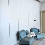 “Two chairs, one perfect corner, zero clutter. Quiet luxury unlocked.” Two modern teal-blue upholstered accent chairs with slim black metal legs positioned side-by-side against a seamless tall white paneled wall. A small round white marble side table between them holds a white vase with soft pink and white floral arrangement, all on light marble flooring in a minimalist interior.