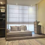 “The reading nook I never knew I needed until right now.” A cozy built-in window seat nook with a low daybed upholstered in beige-and-white gingham check fabric, layered with neutral throw pillows and a folded blanket. The seat is raised on a wooden platform in front of a large window covered by light-filtering beige Roman shades. Flanking the nook are built-in dark-wood shelving on the left and a tall woven-front dresser with a slim black floor lamp on the right, all on light wood-look flooring with a small vintage-style area rug.