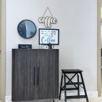 A charming coffee nook blending warm wood textures, playful signage, and minimalist styling. Coffee station with dark wood cabinet, black stool, wall clock, framed quote, and decorative signs.