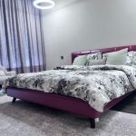 A bold bedroom composition blending vibrant color, minimalist symmetry, and layered material contrast. Modern bedroom with magenta bed frame, green and gray pillows, gray curtains, and circular ceiling light.