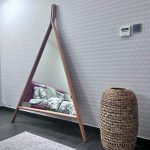 A cozy corner vignette blending geometric reflection, natural textures, and layered interior styling. Triangular wooden mirror leaning against patterned wall, woven basket, dark wood floor, and light rug.