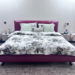 A modern bedroom ensemble blending botanical softness, rich upholstery, and clean-lined symmetry. Maroon upholstered bed with floral bedding, green and gray pillows, minimalist nightstands, and dark wood flooring.