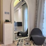A sculptural interior vignette blending reflective geometry, organic textures, and minimalist elegance. Arched mirror with sculptural chair, pedestal with plant, patterned rug, and light wood flooring.