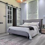 A refined bedroom composition blending ambient lighting, natural textures, and modern architectural accents. Grey upholstered bed with white and grey bedding, textured backlit wall panel, wooden nightstand, and barn-style glass doors.