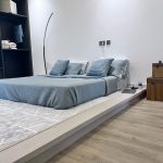 A minimalist bedroom layout blending soft textures, open storage, and functional modern styling. Low-profile bed with light blue bedding, black open wardrobe, wooden nightstand, and geometric area rug.