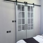 “When your bedroom doors are prettier than most windows.” A pair of light-gray French-style sliding barn doors with upper glass panes divided into six sections each, mounted on a matte-black industrial track. The doors are partially open, revealing a bathroom beyond, positioned at the foot of a neatly made bed in a minimalist bedroom.