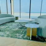 “Twin curved aqua clouds + ocean horizon = the ultimate chill zone.” A luxurious high-floor living room with floor-to-ceiling ocean-view windows. Two identical large, organically curved light aqua-blue plush sofas face each other across a green-abstract area rug. Between them: a round lilac-veined white marble coffee table on a cylindrical base and a smaller round marble side table with a brushed-brass pedestal.