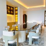 “When your dining table can seat 12 and still look this impossibly chic.” A luxurious modern dining room featuring a long oval black-glass-top table with a dramatic fluted travertine pedestal base, surrounded by twelve sculptural cream bouclé dining chairs with curved wooden frames. Warm LED cove lighting and a backlit onyx bar niche create an elegant golden glow.