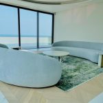 “Curved ice-blue perfection + endless sea views = penthouse goals unlocked.” A luxurious high-floor living room with panoramic sea views through floor-to-ceiling windows. Two large, organically curved light-blue/gray plush sofas face each other around a round white marble coffee table with a sculptural gold base, all placed on a green-abstract area rug over herringbone wood flooring.